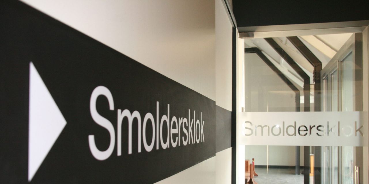 Binnenzicht op de ingang van de Smoldersklok. Aan de muur het strakke zwart-wit logo, in de gang een glazen deur met opnieuw het logo in melkglas.