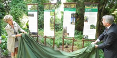 Een vrouw (links) en een man (rechts) houden een groene doek vast, ze staan lachend bij vier banners aan een groene plantenborder in Arboretum Kalmthout, onthulling van infoborden bij de boomplanting.