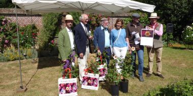 Vijf mannen en een vrouw poseren op een zonnige dag in een tuin voor een aantal rozenstruiken in pot, een van hen houdt fier een certificaat voor met een afbeelding van een roos, doop van een roos tijdens de Tuin- en Plantendagen van Hex 2017.