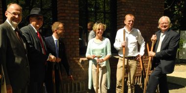 Een plechtig moment bij een boomplanting in Arboretum Kalmthout, 5 mannen en 1 vrouw poseren met een schop in de hand voor de gevel van een gebouw en bij een net aangeplante stek van een druivelaar.