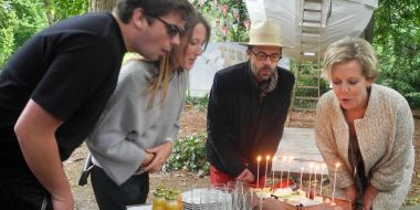 Een jong koppeltje en een man en vrouw blazen tien kaarsjes op een taart uit, op de achtergrond een bolvormige tent in een park, er hangen vlaggenslingers op. Viering 10 jaar Terra Nova in Arboretum Kalmthout.