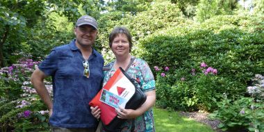 Een man en een vrouw poseren lachend naast bloeiende borders in de tuin van Arboretum Kalmthout. De vrouw houdt een schoudertas vast en toont een ticket waar Terra Nova op staat.